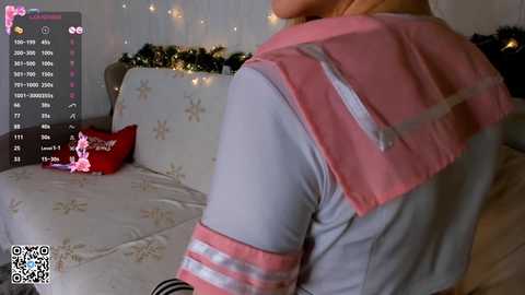 Media: A video of a woman in a pink and white sailor-style top, sitting on a bed with a red heart pillow and floral bedding, surrounded by festive string lights.