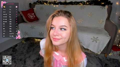 Media: Video of a smiling, fair-skinned, blonde-haired woman in a pink sweater, sitting on a grey carpet, with a festive Christmas-themed couch in the background.