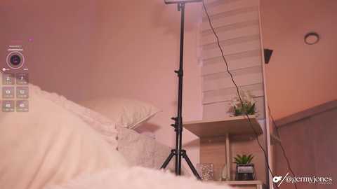 A video of a cozy, pastel-pink bedroom with a white bed, a tall light stand, a white shelf, and a small plant. The background features a wooden staircase and a wall-mounted lamp.