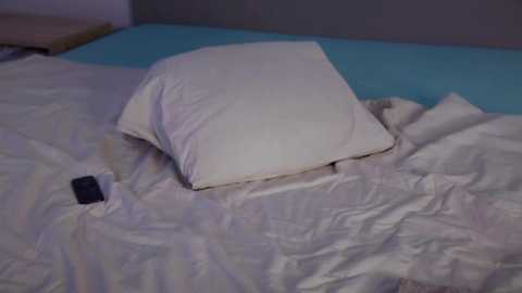 Media: A video of a messy bed with a white pillow, a white duvet, and a blue pillowcase. A smartphone lies on the bed, with a wooden nightstand visible in the background.