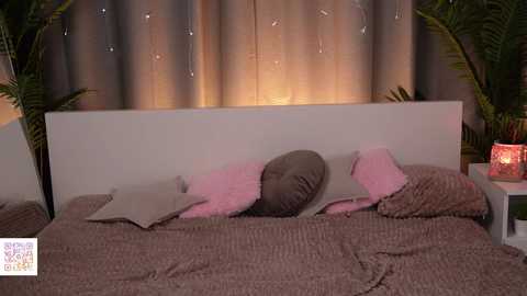 Media: Video of a cozy, dimly lit bedroom with a white headboard, pink and gray pillows, and textured brown blanket on a bed. Two potted plants flank the headboard, and a red-lit candle on a nightstand adds warmth.