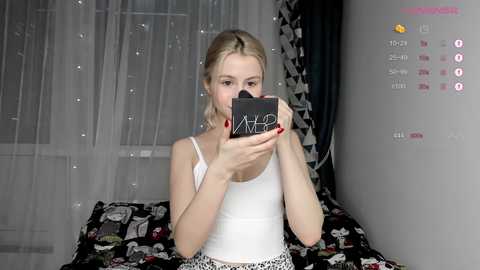 Media: Video of a blonde woman in a white tank top, taking a selfie with a black compact mirror, against a backdrop of a bed with a patterned duvet and white curtains.