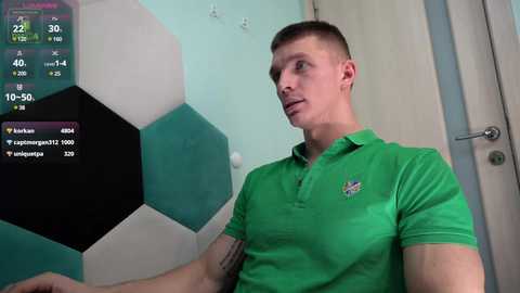 Media: Video of a young, fit man with short hair in a green polo shirt, standing in front of a soccer ball patterned wall, with a digital soccer game display in the top left.