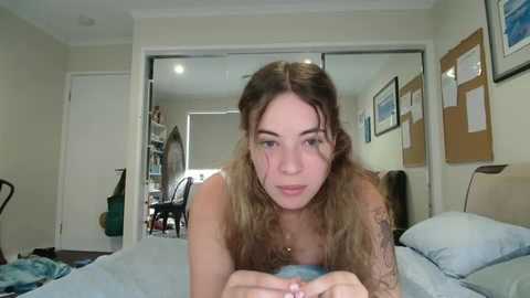 Media: A young woman with long, wavy brown hair, fair skin, and small breasts lies on a bed, looking at the camera. The room has beige walls, a wooden chair, and framed art.