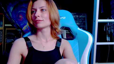 Video of a light-skinned woman with shoulder-length auburn hair, wearing a black sleeveless top, seated in a blue gaming chair, with a dark, cluttered room background.