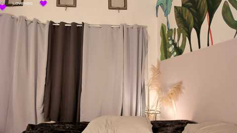 Media: Video of a cozy bedroom with beige walls, green palm tree mural, light gray curtains, potted faux plants, and a warm lamp on a bedside table.