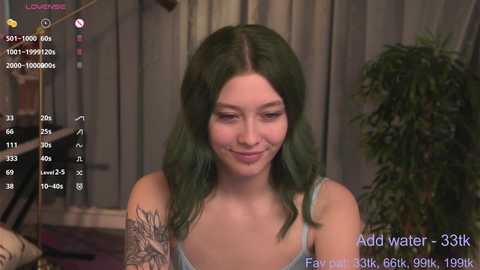 Media: A video shows a young woman with green hair, wearing a light blue tank top, smiling. She has a tattoo on her left arm. The background features a plant and a window. The image is from an OnlyFans account, displaying a live video.