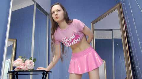 Media: Video of a young Caucasian woman with long brown hair, wearing a pink crop top and matching short skirt, standing in a room with blue walls, floral arrangement on a table, and large mirrors.