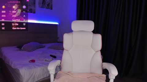 Media: A video of a modern, minimalist bedroom with a white gaming chair in the foreground, a TV displaying a game interface, and a neatly made bed in the background.