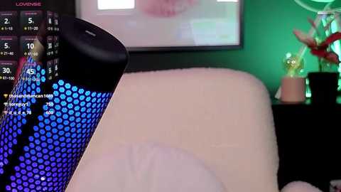 Media: Video of a close-up of a black, glowing, blue LED light bulb on a bed, with a green wall and a partially visible TV screen displaying a makeup tutorial.