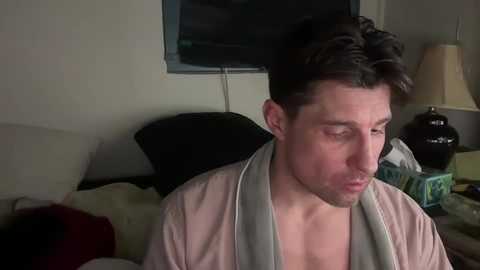 Media: Video of a young man with short brown hair, wearing a light pink robe, sitting on a bed with a dark pillow and beige sheets.