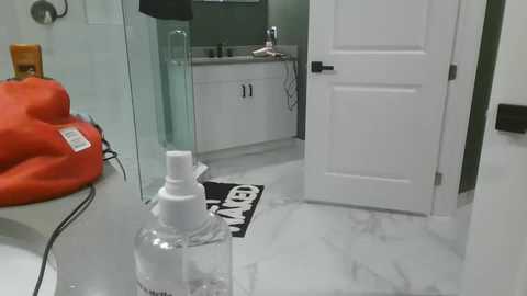 Media: Video of a sleek, modern bathroom with a white marble countertop, a glass shower door, a white door, and a bright orange towel on a white shelf.