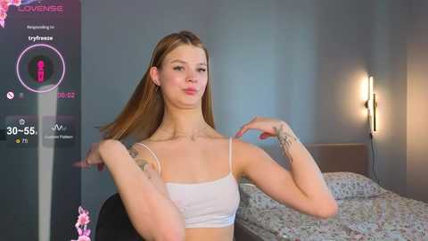 Media: Video of a young woman with fair skin, straight brown hair, wearing a white crop top, sitting on a bed in a modern room with a floral-patterned bedspread and a bedside lamp.