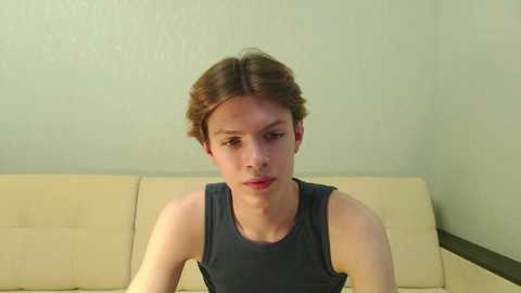 Media: Video of a young, Caucasian boy with shoulder-length brown hair, wearing a dark gray tank top, sitting on a beige leather couch against a light green wall.