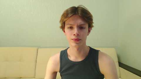 Media: Video of a young, fair-skinned, androgynous person with shoulder-length brown hair, wearing a dark gray sleeveless shirt, seated on a beige couch against a plain green wall.