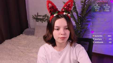 Media: Video of a young woman with light skin, shoulder-length dark hair, wearing a festive red headband with a bow and white rose. She sits on a beige bed with a fluffy blanket, in a cozy room with a potted plant and purple lighting.