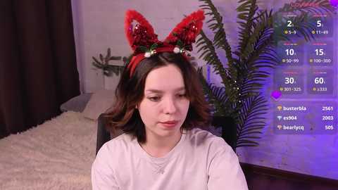 Media: Video of a young woman with fair skin and shoulder-length brown hair, wearing a red, furry rabbit headband. She sits in a plush, white chair, surrounded by a cozy, dimly lit room with a large, green fern plant in the background.