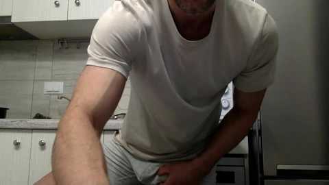 Media: Video of a muscular man in a white T-shirt and gray shorts, leaning over a kitchen sink with white cabinets and beige tiles in the background.