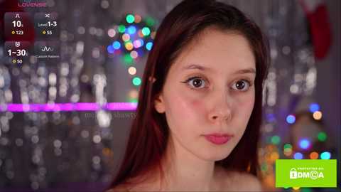 Media: Video of a young woman with fair skin and long brown hair, wearing a pink shirt, standing in front of a blurred, colorful, festive background. Overlay includes streaming platform logo and chat details.