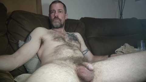 Media: Video of a bald, bearded, white man with a muscular build, lying naked on a brown couch. He has a large, circumcised penis and is positioned with legs spread.