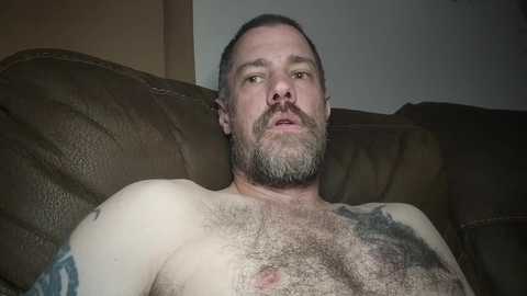 Media: Video of a muscular, middle-aged Caucasian man with short hair and a beard, lying shirtless on a brown leather couch. He has a tattoo on his right arm and a scar on his chest. Background is plain with dim lighting.