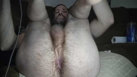 Media: Video of a nude, overweight man with a hairy chest and pubic area, lying on a beige couch with legs raised, revealing his genitals and anus. Background shows a dark brown couch and a blue water bottle.