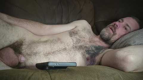 Media: Video of a nude, bearded Caucasian man with a hairy chest and a circumcised penis, lying on a beige couch, holding a black remote control.