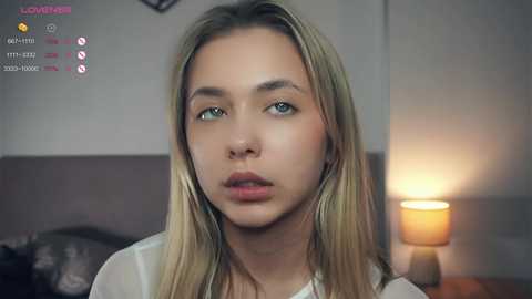 Media: Video of a young woman with long blonde hair, blue eyes, and light skin, wearing a white shirt. She has a neutral expression. Background features a lamp and dark furniture.