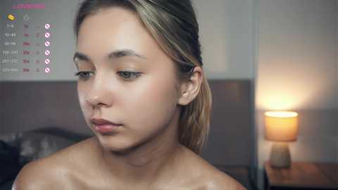 Media: Video of a young, light-skinned woman with light brown hair, looking introspective, in a dimly lit bedroom with a lamp and calendar.