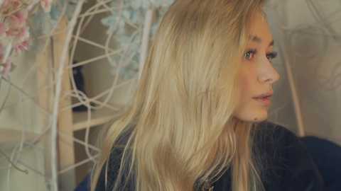 Media: Video of a young, light-skinned blonde woman with long, straight hair, wearing a black leather jacket, in a softly lit room with pastel floral decor and a white geometric mirror in the background.