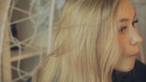 Media: Video of a blonde woman with straight, shoulder-length hair, viewed from the side. She has light skin, a slight smile, and wears a black top. Background features a white, geometrically-patterned wall.