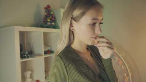 Media: Video of a young, fair-skinned woman with blonde hair in a green ribbed cardigan, looking contemplative in a softly lit room with a white shelving unit filled with decorative items.