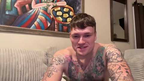 Media: Video of a shirtless, tattooed man with short dark hair, smiling, in a living room with a colorful, abstract painting and a framed mirror.