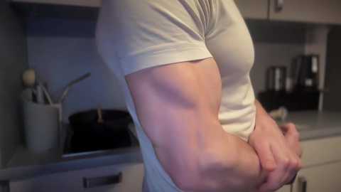 Media: Video of a muscular man in a tight white t-shirt, showcasing defined biceps and triceps, standing in a modern kitchen with stainless steel appliances, black pans, and utensils in a white holder.