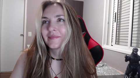 Media: Video of a smiling, fair-skinned woman with long, straight blonde hair, wearing a black choker and a red and black gaming chair in a modern, minimalist room with white walls, a window, and a small action figure.