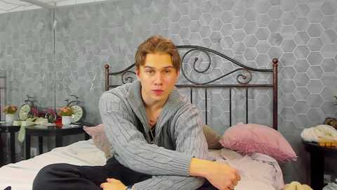 Media: Video of a young man with fair skin, brown hair, wearing a gray sweater, lying on a bed with a black metal headboard in a gray patterned room.