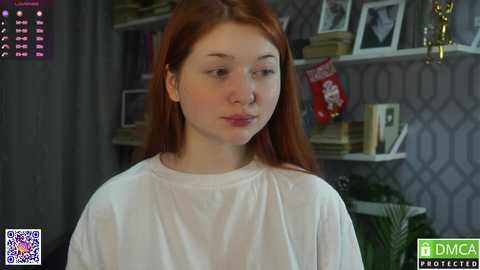 A video of a young woman with long, straight red hair, fair skin, and a slender build, wearing a white t-shirt, standing in a room with a white wall, bookshelves, and a Christmas decoration.