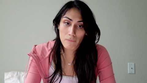 Media: Video of a young woman with long, straight black hair, wearing a pink jacket over a white tank top, standing against a plain, light-colored wall.