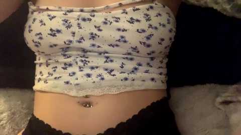 Media: Video of a light-skinned woman with a flat stomach, wearing a white strapless crop top with blue floral patterns and black lace-trimmed panties, lying on a bed with a beige blanket.