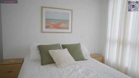 Media: A video of a minimalist bedroom with a white bed, two green and one white pillow, and a framed painting of a beach on the wall. Light streams through sheer white curtains.