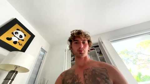 Media: A video of a shirtless young man with curly hair and tattoos on his chest, standing in a bright room with a panda-themed lamp, white walls, and large windows.