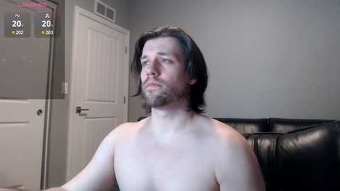 Media: Video of a shirtless man with shoulder-length brown hair, fair skin, and a trimmed beard, sitting on a black leather couch in a dimly lit room with gray walls and a white door.