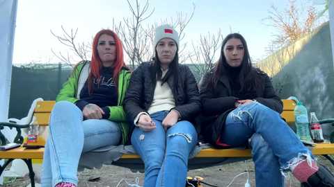 Media: Video of three young women sitting on a yellow bench, wearing casual clothing, with bare trees in the background.