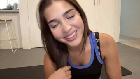 Media: Video of a smiling young woman with straight, dark brown hair, wearing a black and blue sports bra, sitting on a black yoga mat in a modern, minimalist room with white walls and gray carpet.