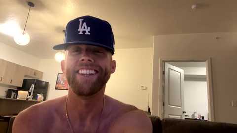 Media: Video of a shirtless, muscular, bearded white man with a trimmed beard, wearing a blue LA Dodgers baseball cap, smiling in a modern, dimly-lit living room with a kitchen in the background.