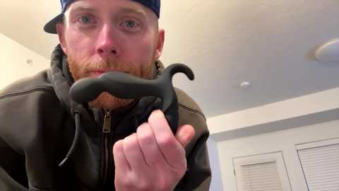 Media: Video of a bearded man with a red beard, wearing a dark jacket and blue baseball cap, holding a black mustache prop close to his face. Background shows white walls and a ceiling light.