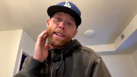 Media: Video of a bearded, fair-skinned man with glasses, wearing a dark jacket, blue LA Dodgers cap, and touching his chin. Background shows a ceiling light and white walls.