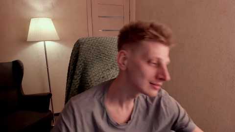 Media: Video of a young man with short brown hair and a light complexion, wearing a gray t-shirt, sitting in a chair with a textured green blanket, in a dimly lit room with beige walls and a lamp.