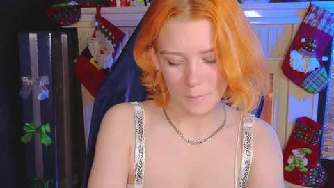 Media: Video of a young woman with orange hair, wearing a white camisole with silver chain necklace, standing in a cozy, festive room with Christmas stockings and gifts.