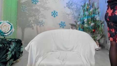 Media: Video of a cozy, modern living room with a white, plush armchair draped in a white blanket. Background features a green wall, a Christmas tree adorned with blue and white ornaments, and a person in a red and black dress standing near the tree.